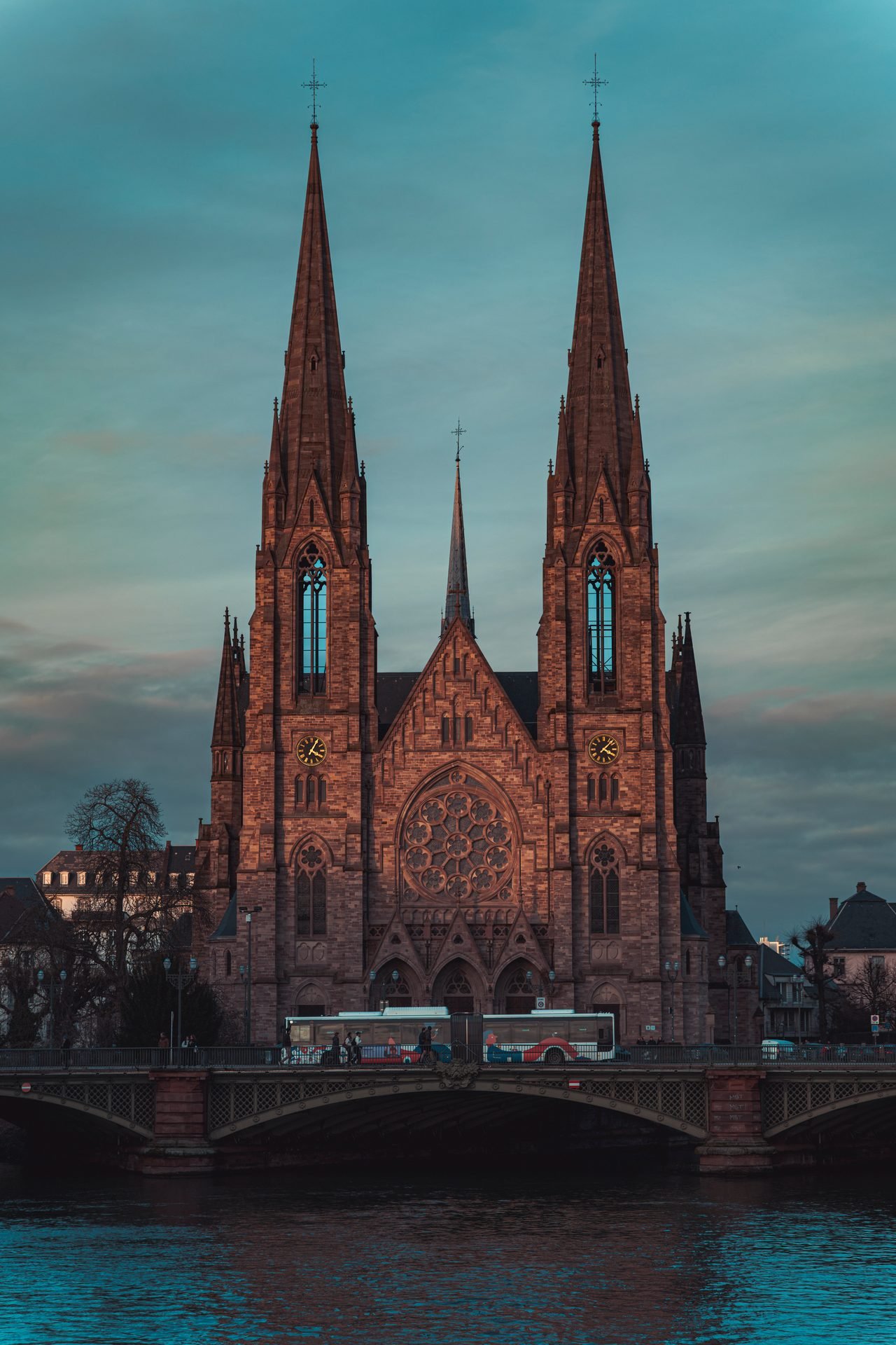 Neugotische Kirche mit zwei Türmen am Flussufer, davor eine Brücke in der blauen Stunde