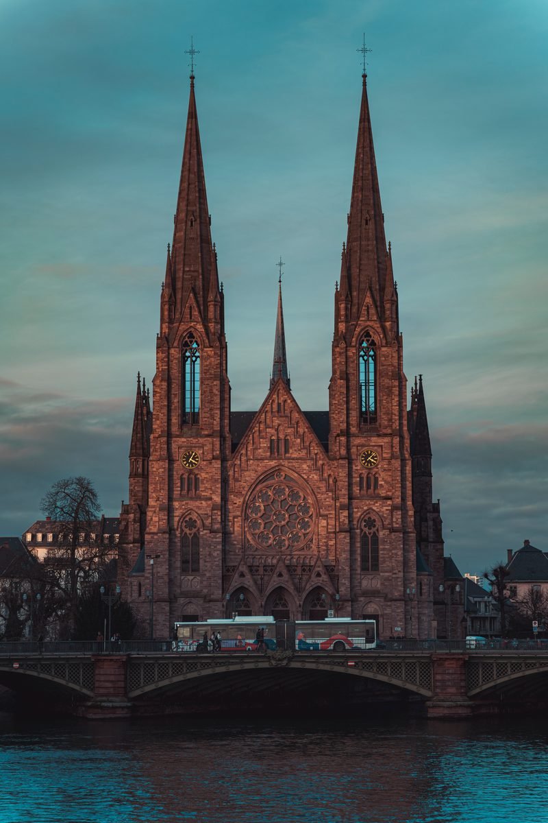 Neugotische Kirche mit zwei Türmen am Flussufer, davor eine Brücke in der blauen Stunde
