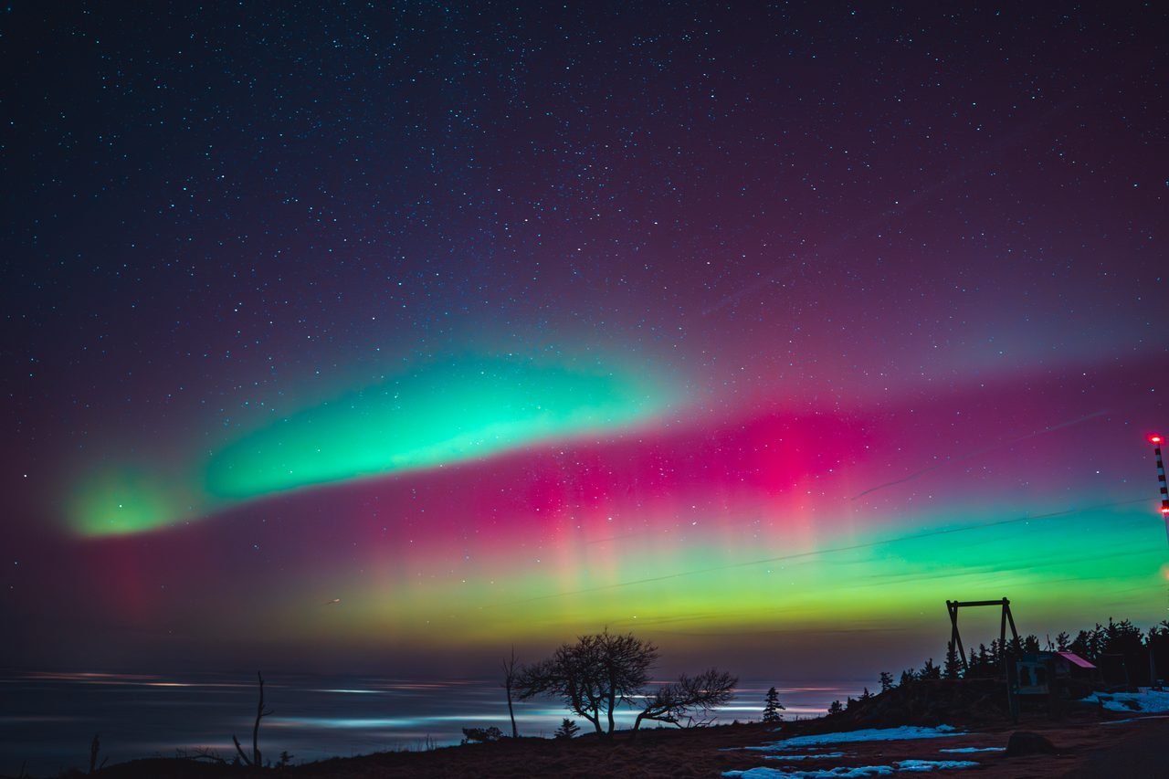 Polarlicht in Grün und Magenta über einer dunklen Landschaft am Wasser