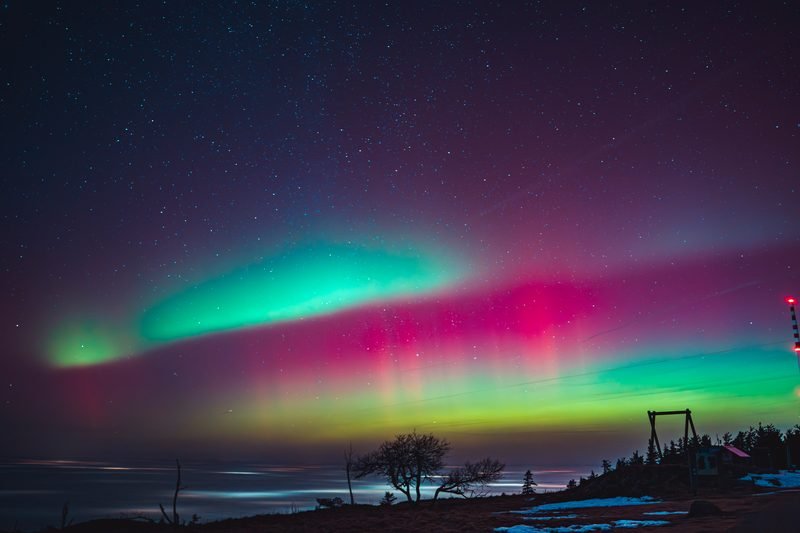 Polarlicht in Grün und Magenta über einer dunklen Landschaft am Wasser