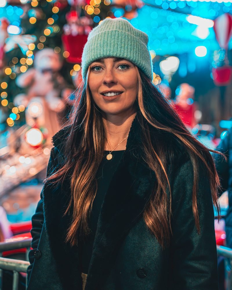 Frau mit mintgrüner Mütze lächelt vor den bunten Lichtern eines Weihnachtsmarkts